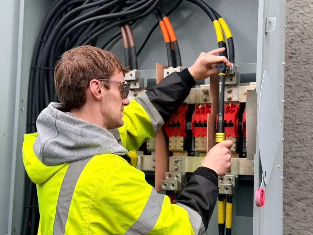 Electrician in high-visibility jacket using a torque wrench to tighten cables inside an industrial electrical panel.
